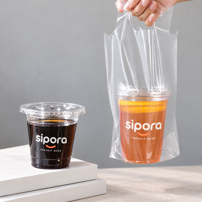 Hand holding a clear single-cup PE carrier bag with iced tea; a coffee cup sits beside it.